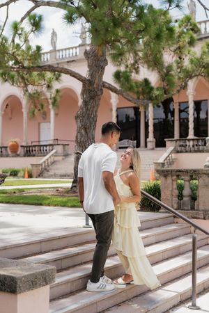 A couple embraces on stone steps outside a pink Mediterranean-style building with arched colonnades.