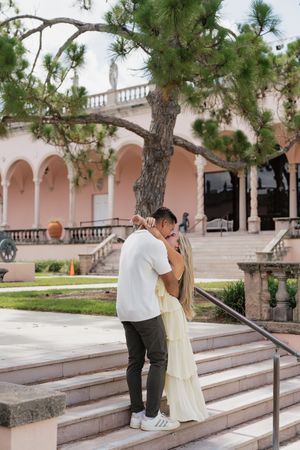 Engaged couple sharing kiss on steps beneath pine tree at pink Mediterranean building.