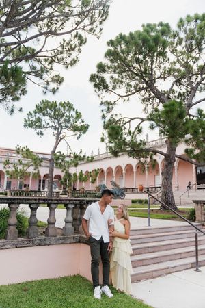 Couple embracing and gazing at each other beside stone balustrade with pink arched colonnade and tall pine trees.