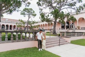A couple poses together on the grounds of a grand pink Mediterranean-style building with arched colonnades.