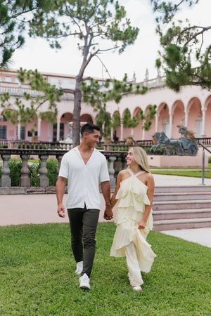 Couple holding hands walking on grass in front of a pink Mediterranean-style building with arches.