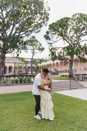 Couple sharing romantic kiss on manicured lawn in front of pink Mediterranean-style building with tall trees.
