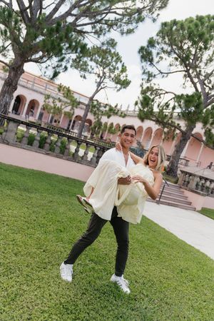 Man carrying laughing woman in his arms on green lawn in front of a pink arched estate.