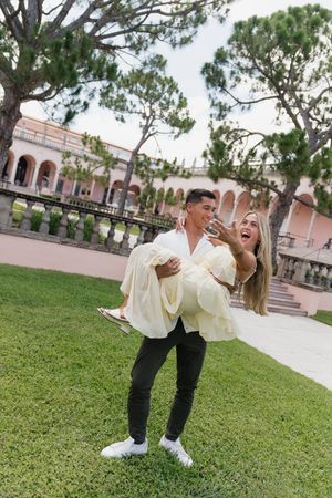 Excited woman being carried by fiance showing off engagement ring outdoors at estate.