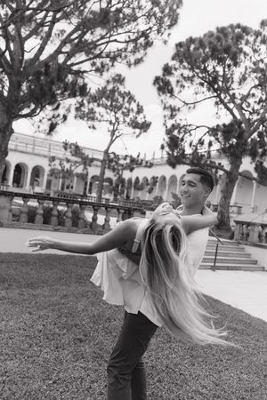 Black and white photo of man joyfully spinning woman with flowing blonde hair in historic garden setting.