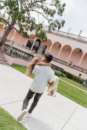 Man lifting woman in front of a pink arched building during an outdoor couples photo session.