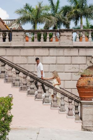 Couple holding hands while descending ornate stone staircase flanked by palm trees at historic estate.