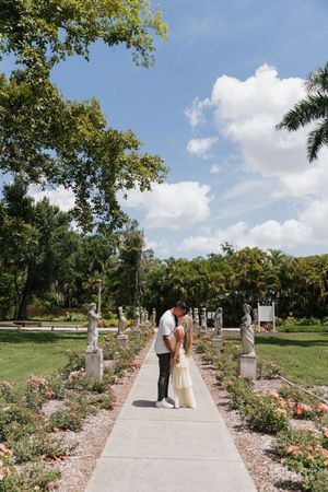 Couple sharing a kiss on a garden pathway lined with statues and roses under a blue sky.