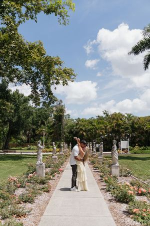 Couple dancing on a garden path lined with statues and roses under a blue sky.