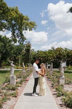 Couple standing face to face on a garden path surrounded by statues and roses.