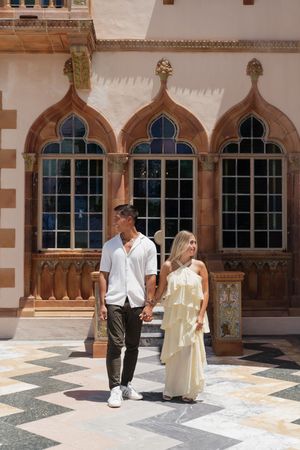 Couple holding hands walking in front of ornate Venetian Gothic architecture with arched windows.