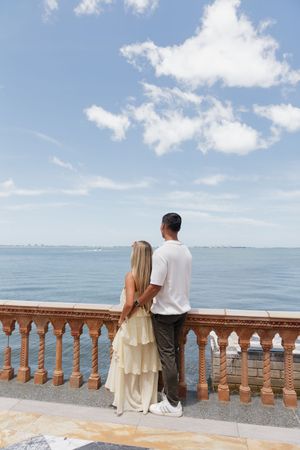 Couple standing together at ornate balustrade overlooking calm blue ocean under partly cloudy sky.