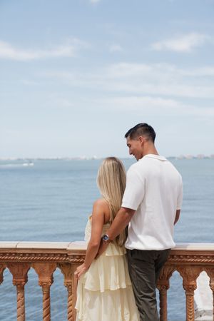 Romantic couple standing on ornate balcony overlooking blue ocean, woman in yellow dress and man in white shirt.