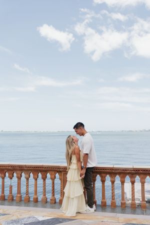 Couple holding hands on ornate terrace overlooking calm blue ocean under cloudy sky, woman in tiered cream dress.