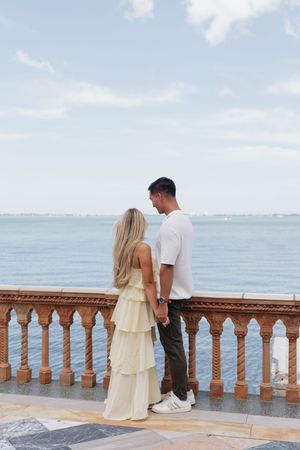 Couple holding hands on ornate balcony overlooking the sea, woman in tiered yellow dress, man in white shirt.