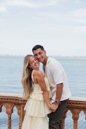 Smiling couple embracing on a scenic ocean balcony, woman in yellow ruffle dress, man in white shirt.
