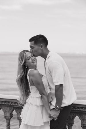Black and white photo of a couple sharing a romantic kiss on a waterfront balcony.