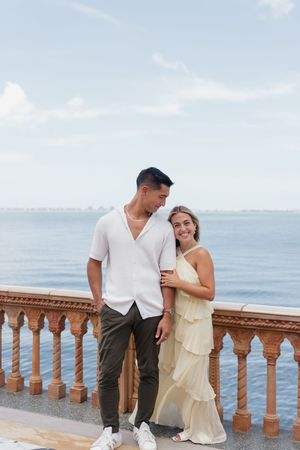 Happy couple standing together on an oceanfront balcony, man in white shirt and olive pants, woman in yellow dress.