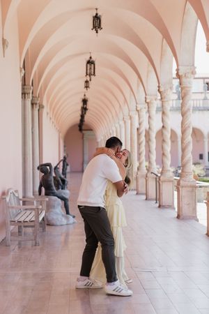 Couple embracing and kissing under elegant pink arched colonnade with bronze sculptures.