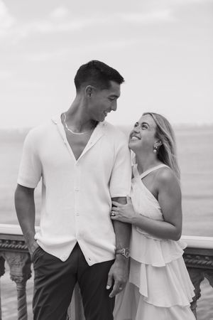 Black and white photo of a smiling couple in white outfits embracing on a seaside balcony.