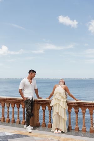 Couple posing on an ornate seaside balustrade, woman in ruffled cream dress, man in white shirt, ocean backdrop.