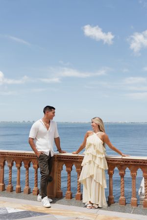 Couple in elegant outfits standing by ornate balustrade overlooking ocean on sunny day.