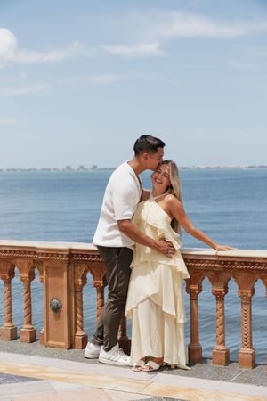 Couple embracing and kissing on a scenic waterfront balustrade overlooking the ocean on a sunny day.