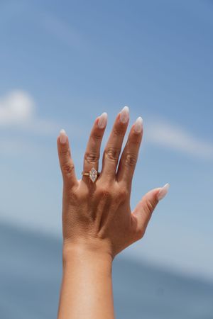 Close-up of a hand raised against blue sky showing a marquise diamond engagement ring.
