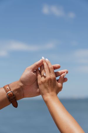 Close-up of hands showing engagement ring against blue sky with ocean in background.