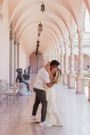 Couple sharing a romantic dip kiss in a pink arched colonnade with sculptures and hanging lanterns.