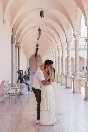 Couple sharing a romantic kiss under elegant arched colonnade with marble columns and bronze sculptures.