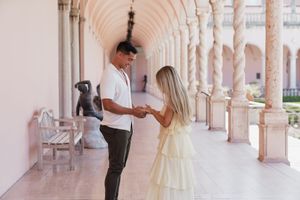 Couple holding hands in elegant pink archway colonnade with marble columns and sculpture.