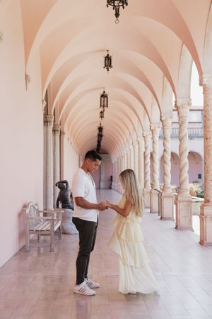 Couple holding hands and gazing at each other in an elegant pink arched corridor with marble columns.