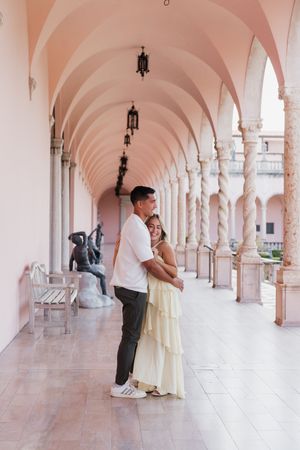 Happy couple hugging under a pink arched colonnade with marble columns and sculptures.