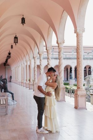 Couple sharing a romantic kiss under pink arched colonnade at Ringling Museum, woman in yellow dress.