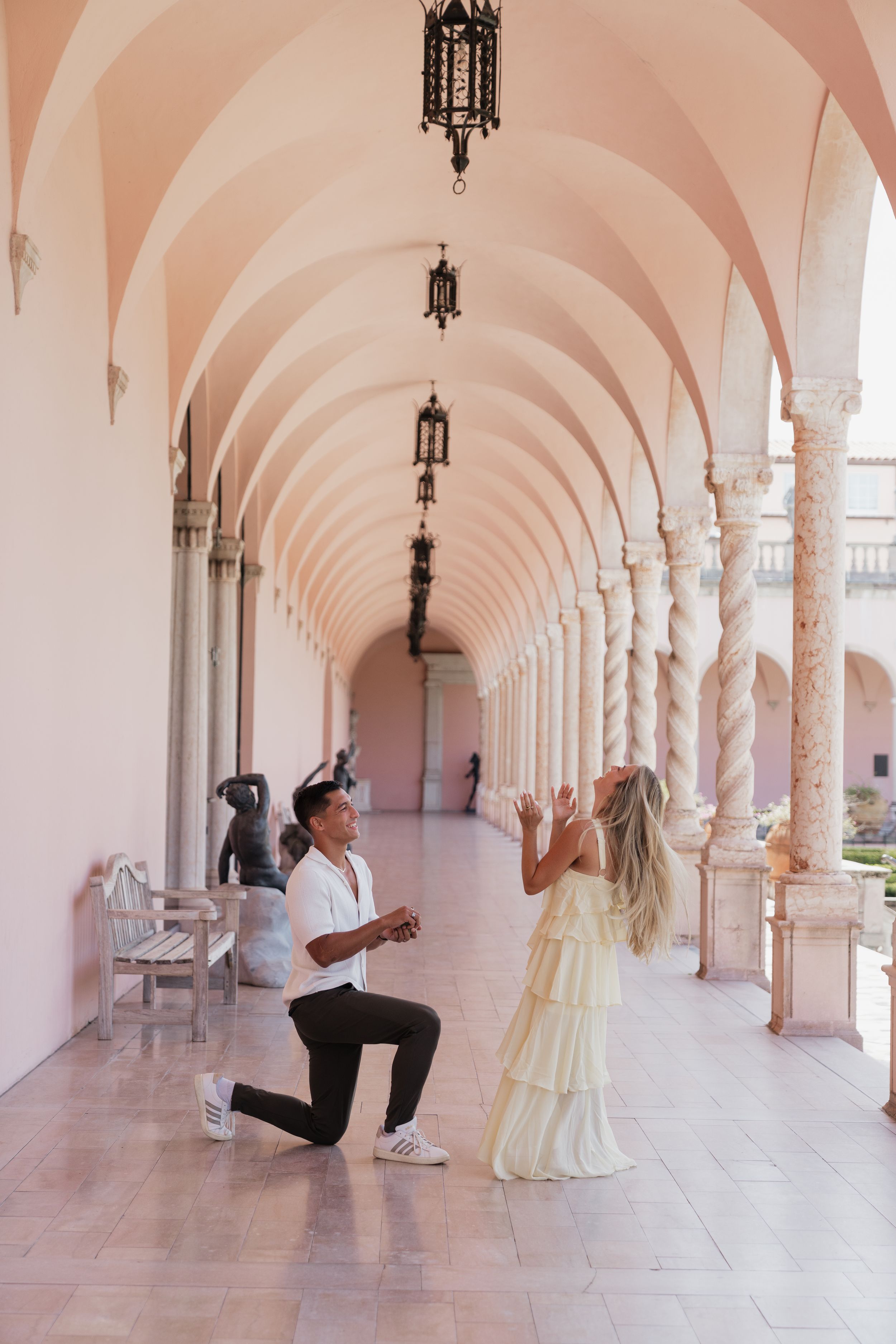 Man on one knee proposing to woman in yellow dress under pink arched colonnade.