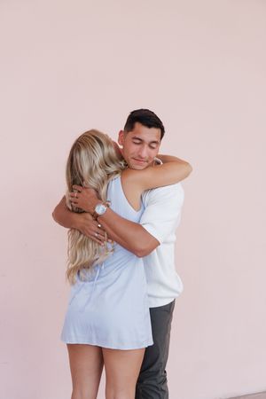 A couple shares a warm embrace against a soft pink wall, the woman in a light blue dress with long blonde hair.