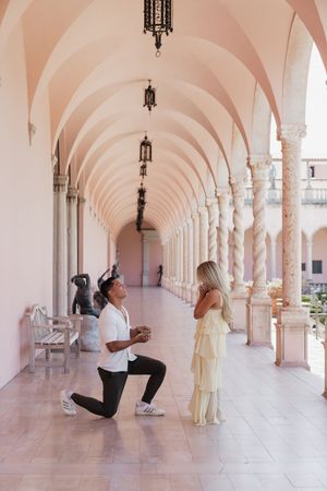 Man proposing on one knee to woman in yellow dress under pink arched colonnade.