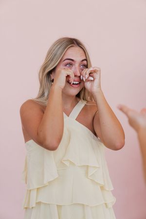 Blonde woman in cream ruffled dress laughing and wiping tears of joy against a pink background.