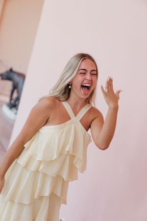 Excited blonde woman in ruffled yellow dress laughing and showing engagement ring on her hand.