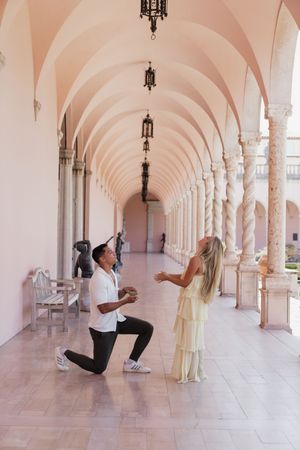 Man proposing on one knee to woman in elegant yellow dress in pink arched colonnade.