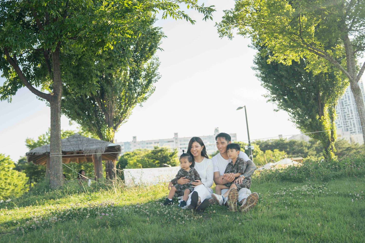 Family of four sitting on lush green grass in a tree-lined park on a sunny day.