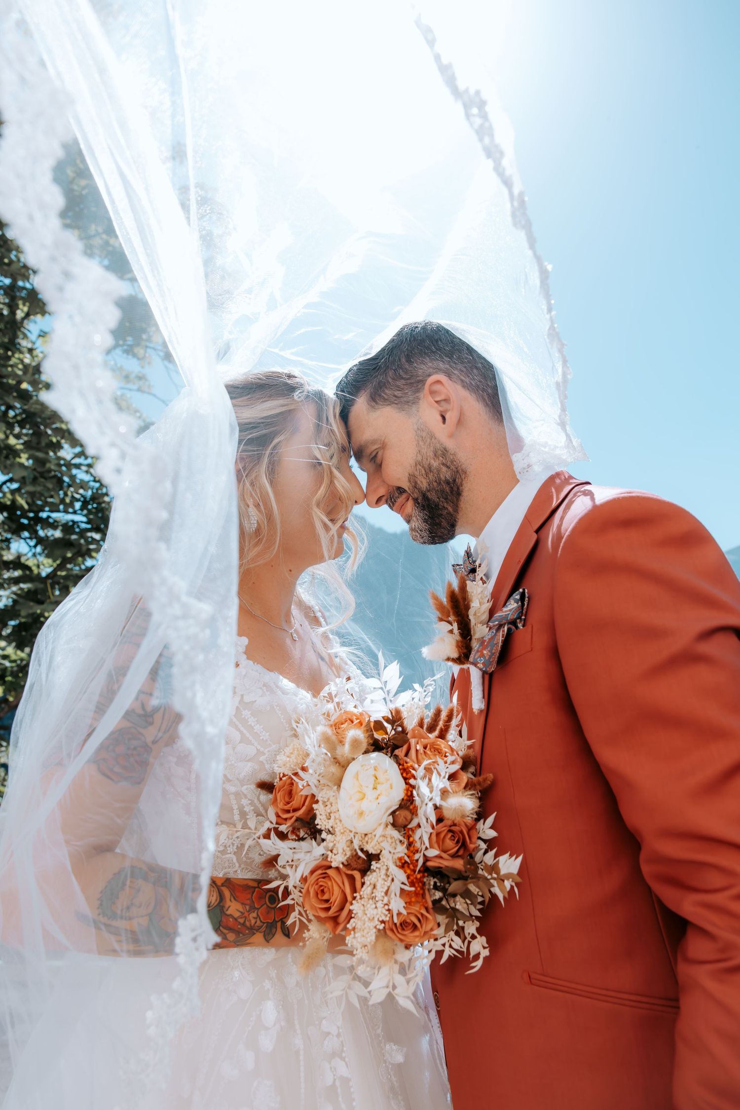 Bride and groom share an intimate moment under a lace veil, holding a boho bouquet with orange and white blooms.