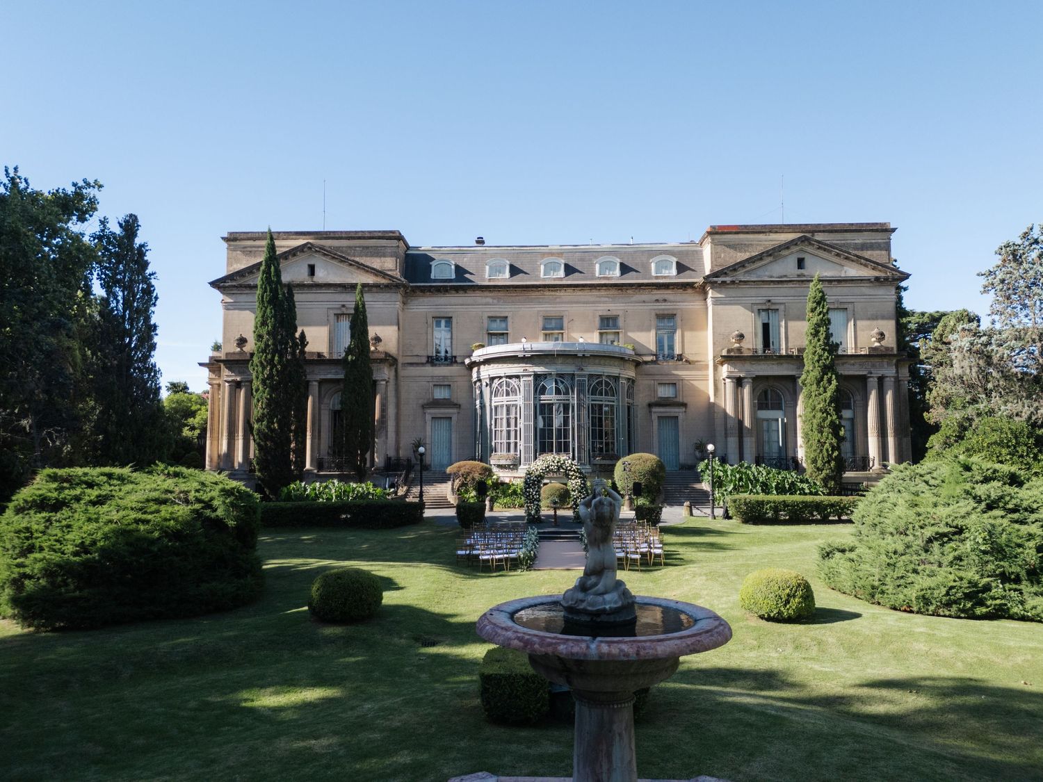 Elegant mansion wedding venue with fountain and manicured gardens under blue sky.
