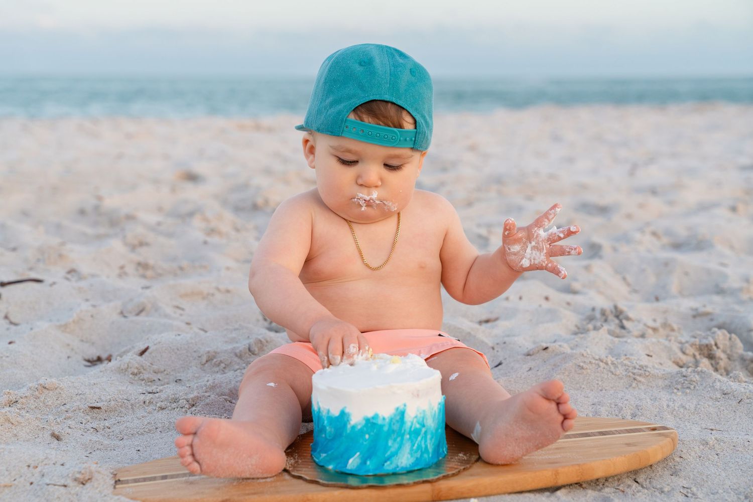 Baby in teal cap enjoys first birthday cake smash on sandy beach, covered in blue and white frosting.