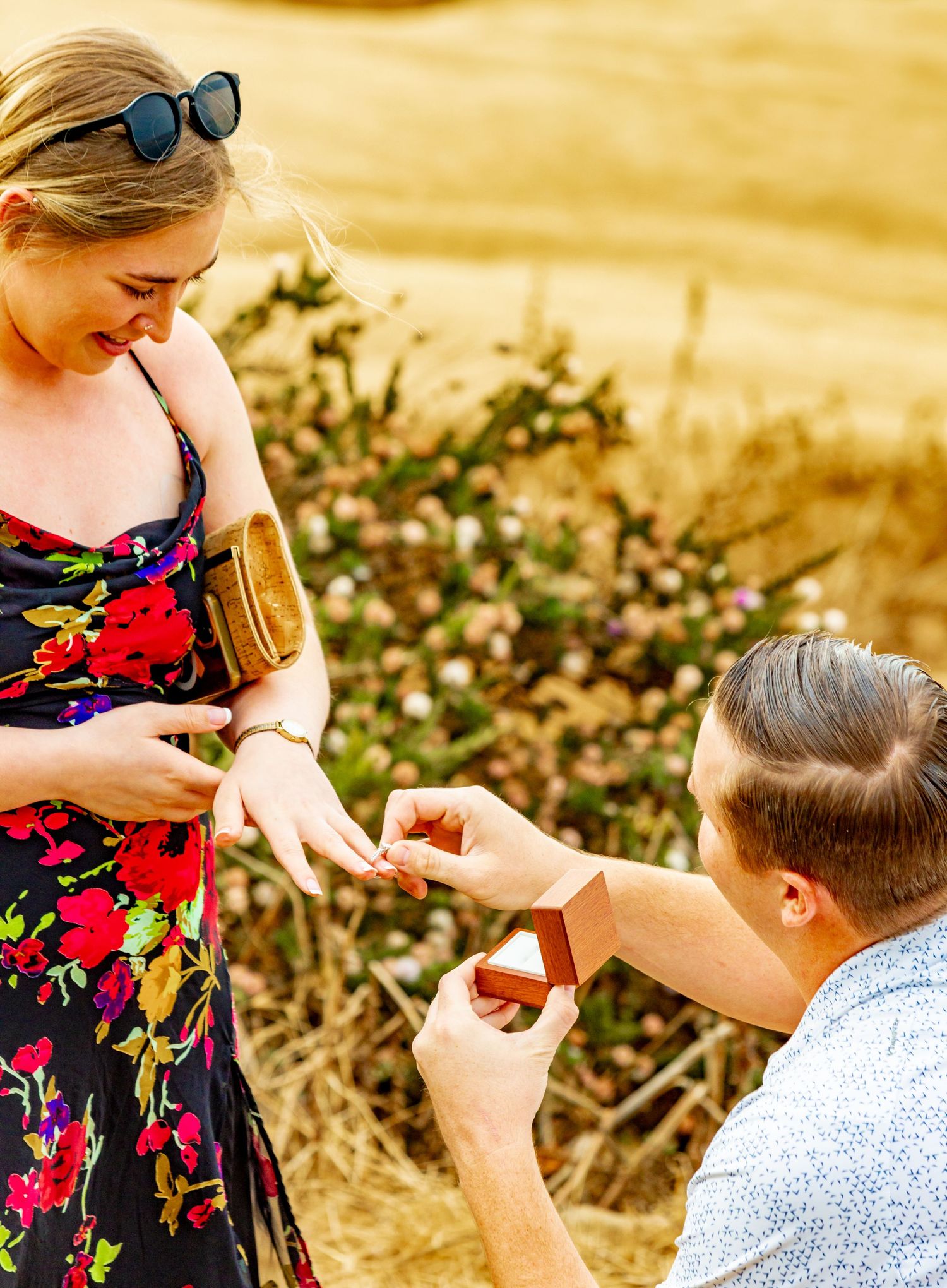 Man kneeling on one knee proposing to a smiling blonde woman outdoors in a golden field with wildflowers.