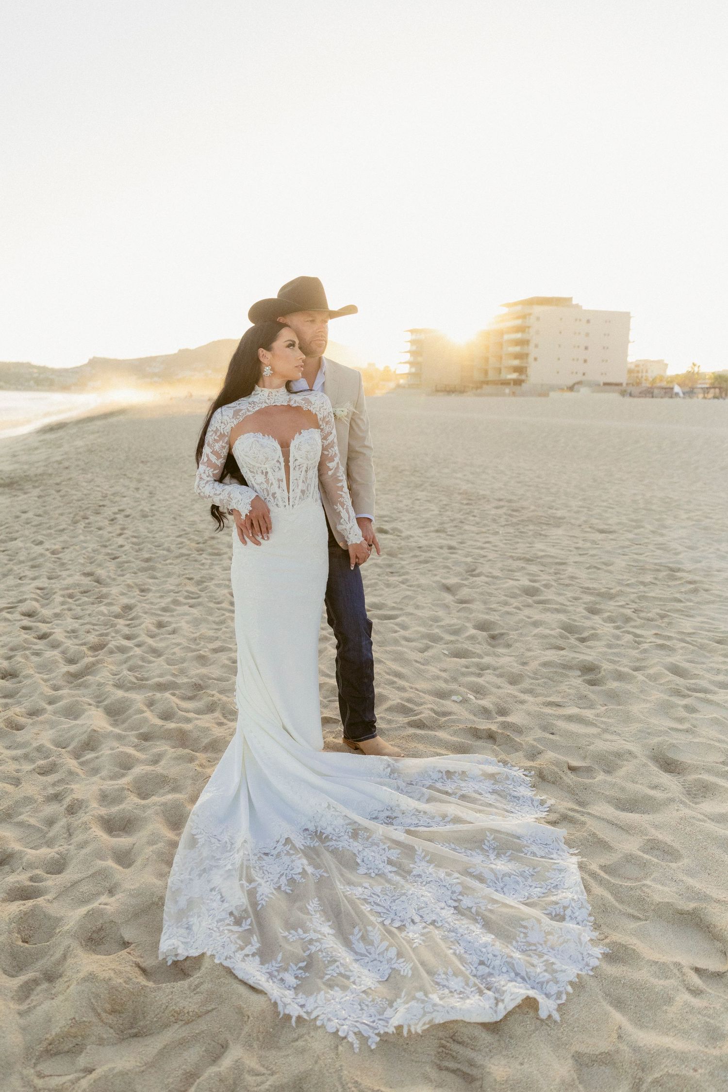 Bride in lace gown and groom in tan suit share sunset beach moment in Cabo.