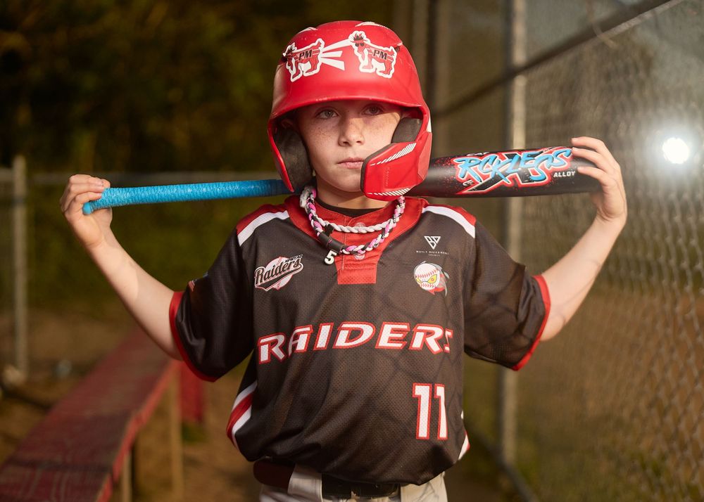 Young baseball player in Raiders jersey number 11 poses with bat behind neck wearing red helmet at night.