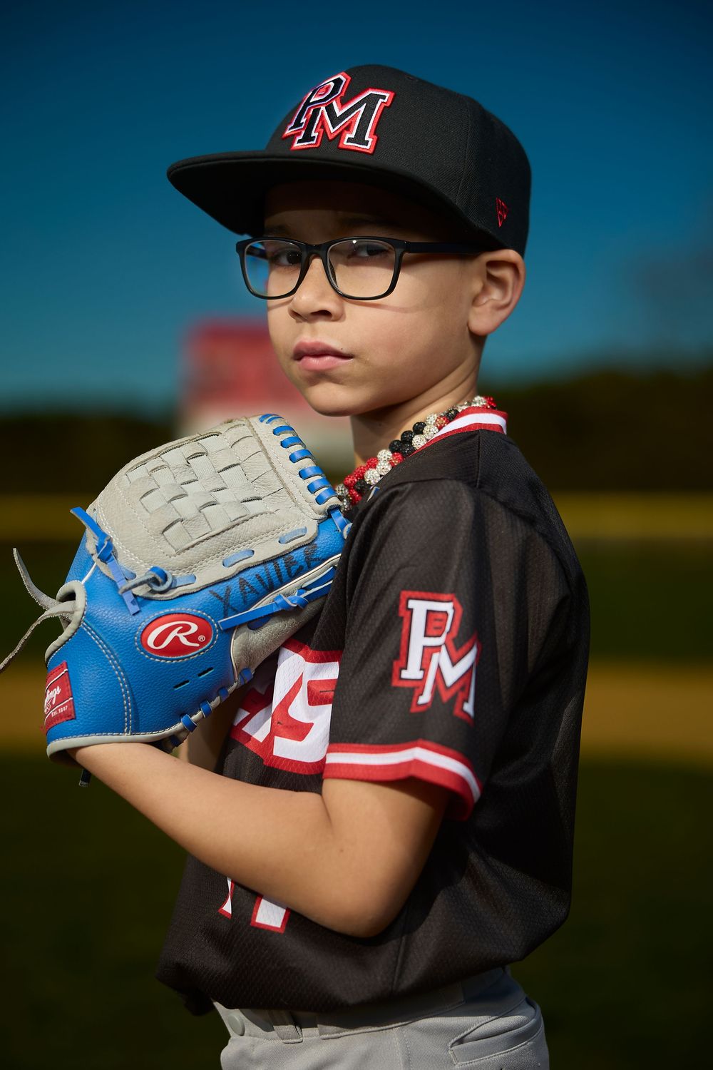 Young baseball player in PM team uniform and cap holds Rawlings glove on field.