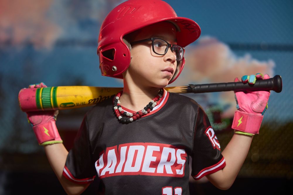 Young baseball player in Raiders uniform and red helmet holds pink bat over shoulders, wearing glasses and pink batting gloves.